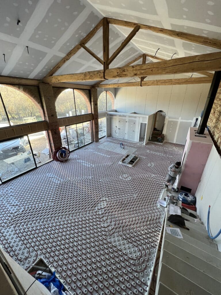Grande pièce en construction : chauffage au sol radiant, poutres apparentes, grandes baies vitrées. Murs en plâtre et pierre. Un ouvrier travaille.