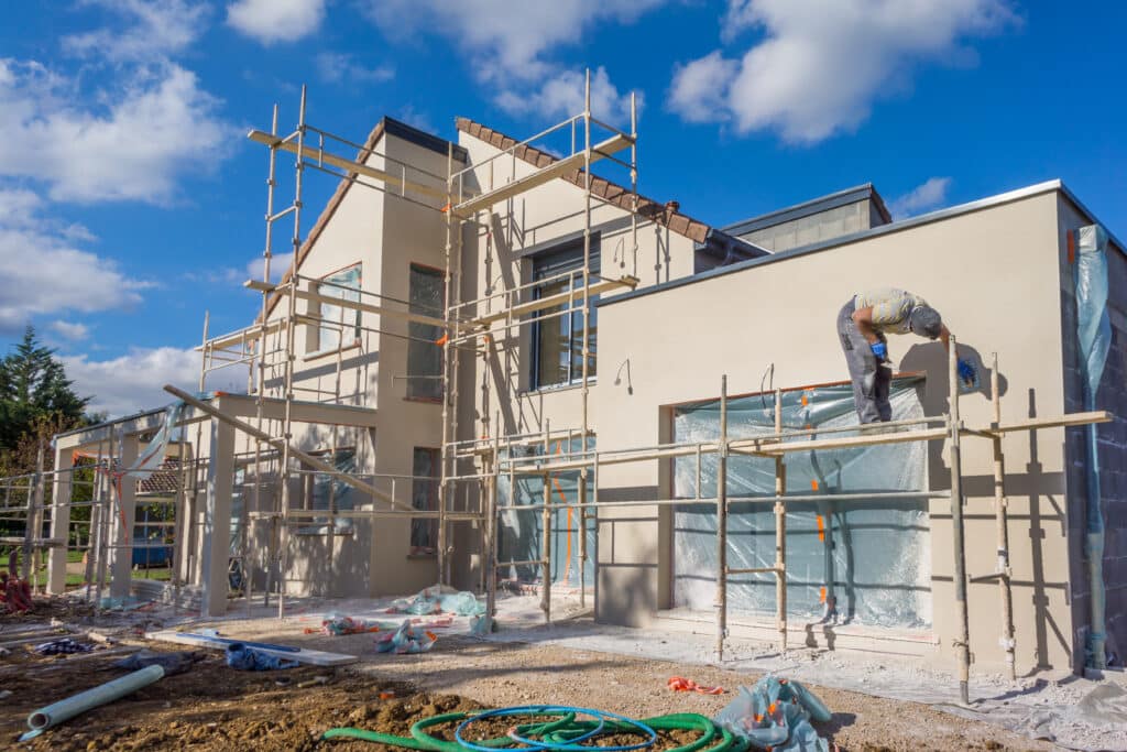 Maison moderne en construction, échafaudage et ouvrier appliquant l'enduit sur la façade claire. Chantier sous ciel bleu ensoleillé.