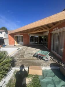 Maison en construction avec patio béton, pergola bois neuve, portes coulissantes et mur TERREAL sous ciel bleu.