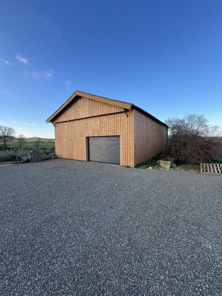 Bâtiment agricole moderne en bois avec bardage vertical et porte de garage gris foncé. Allée en gravier, paysages vallonnés et ciel bleu.