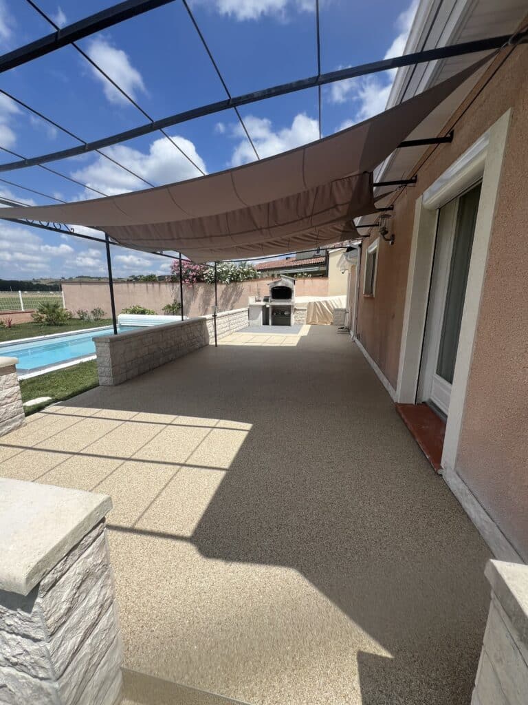 Patio avec pergola ombragée, sol en résine, piscine bleue et barbecue en pierre. Vue sur un paysage verdoyant sous ciel nuageux.