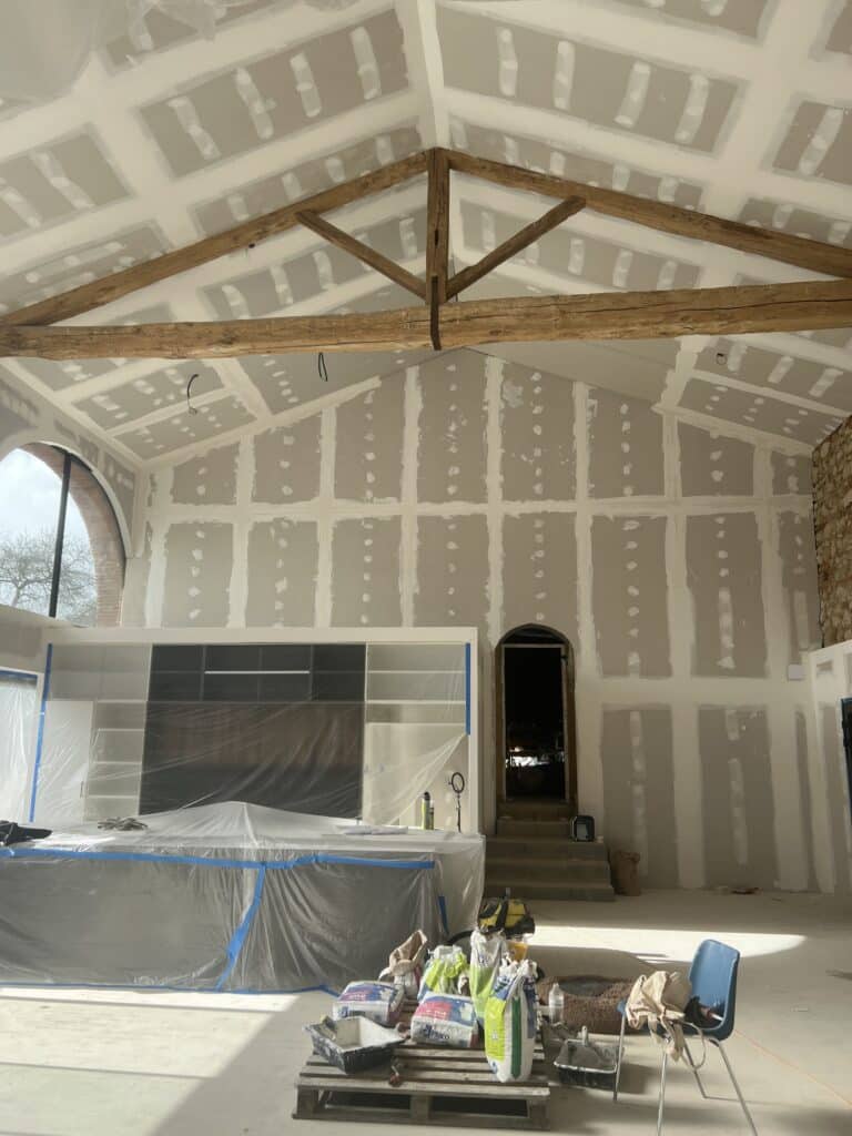 Grande pièce en rénovation avec plafond voûté, poutres apparentes et murs en placo brut. Cuisine bâchée et matériaux 'Placo'.