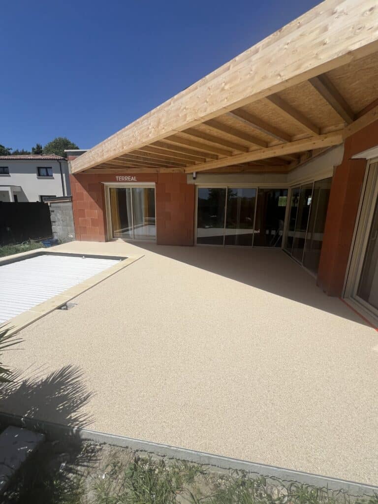 Grande terrasse en résine sous une pergola bois, façade brique TERREAL, piscine couverte. Maison en construction.
