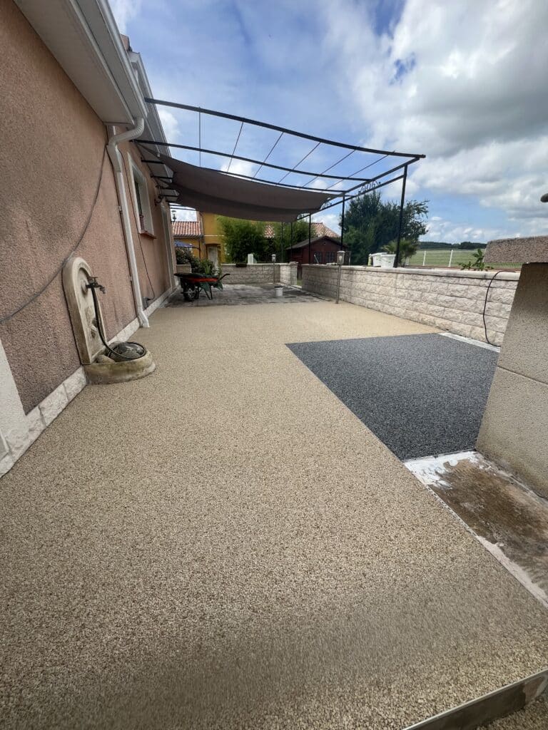Terrasse moderne en graviers de résine beige et gris, adossée à une maison rose avec pergola et aménagement de jardin.