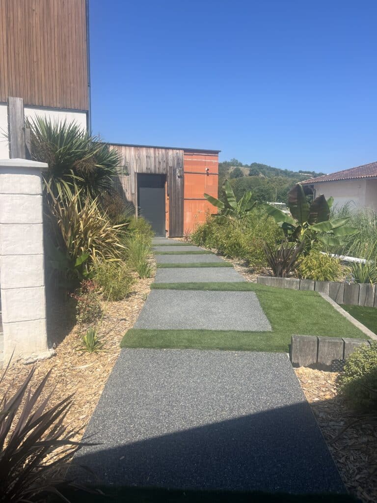 Allée de jardin moderne avec chemin en dalles de gravier et gazon synthétique. Plantes exotiques, paillis. Façade bois et briques, ciel bleu.