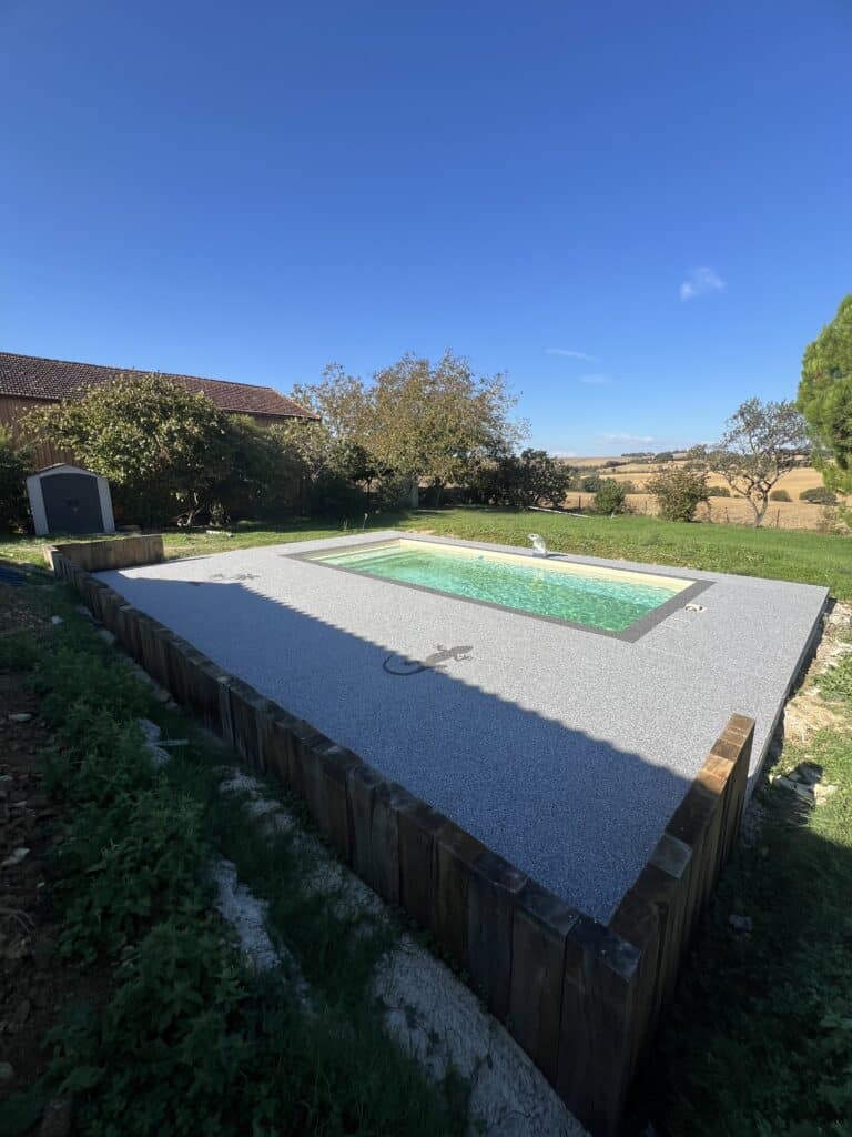 Petite piscine rectangulaire moderne sur terrasse mouchetée grise avec motif gecko, bordée de bois. Arrière-plan champêtre sous ciel bleu.