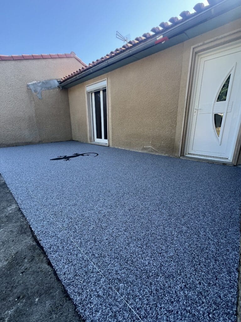 Nouveau revêtement de sol extérieur bleu-gris en résine avec motif gecko noir, devant une maison beige à toit tuilé, sous ciel bleu.